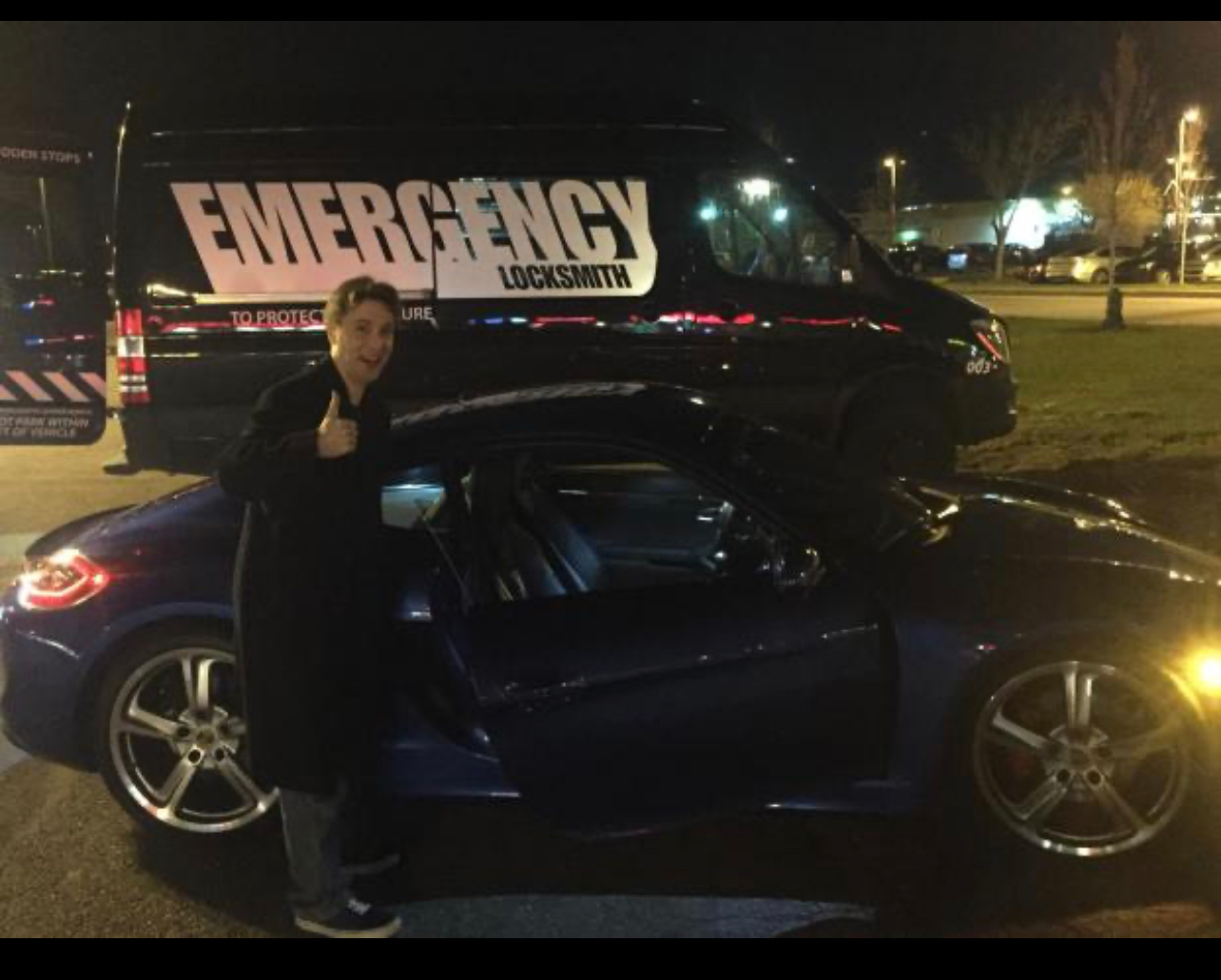 Locksmith giving a thumbs-up beside a blue car and branded service van at night